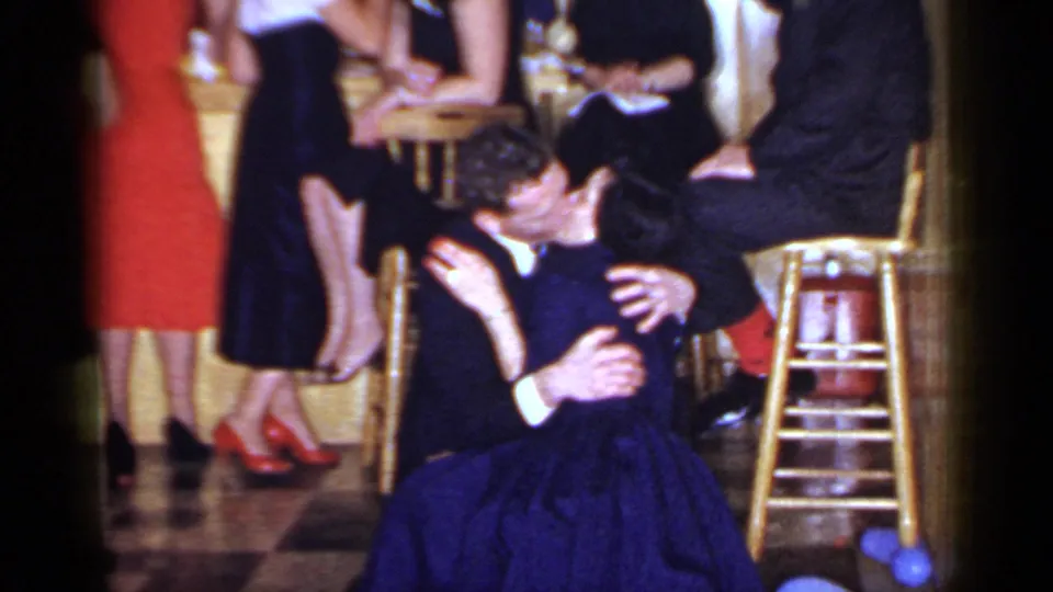 1966 Mineola home-movie frame of a couple dancing closely near bar stools, with formal guests and a red dress visible behind them.