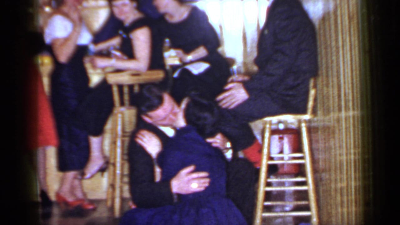 A suited man embraces and kisses a woman in a blue dress beside guests and wooden bar stools.