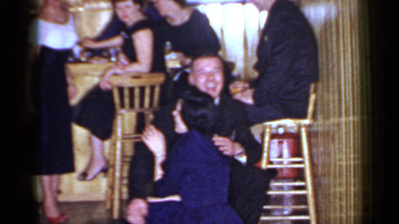 A man smiles while holding a woman in a blue dress near seated guests and stools.