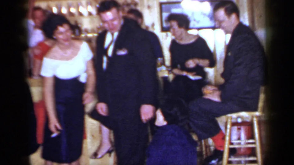 1966 Mineola home-movie frame showing adults in formal dress around a couple on a checkered floor inside a private-looking room.