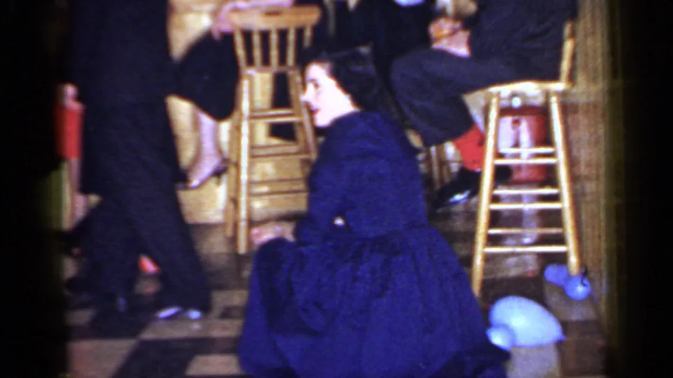 1966 Mineola indoor social frame showing a woman in a blue dress low to the floor near bar stools and guests' legs.