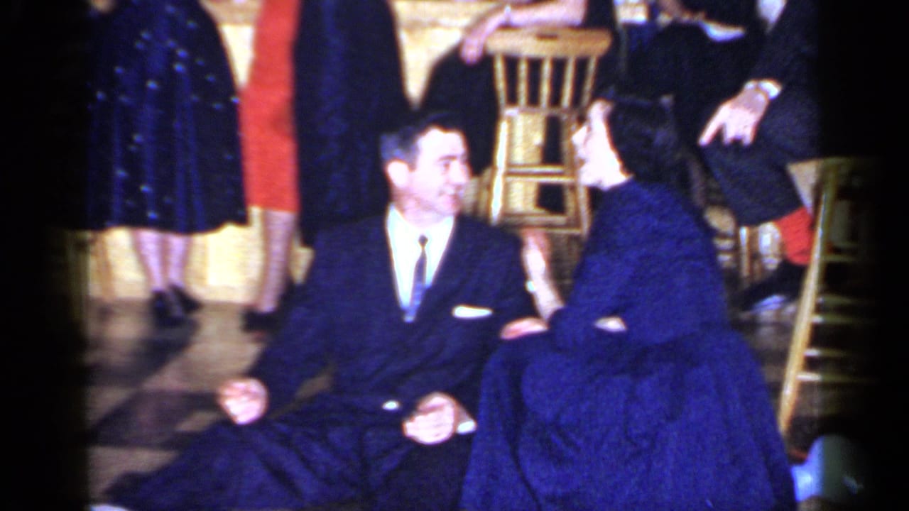 The couple sits close together on the Mineola dance floor, surrounded by guests in formal clothing and wooden stools.