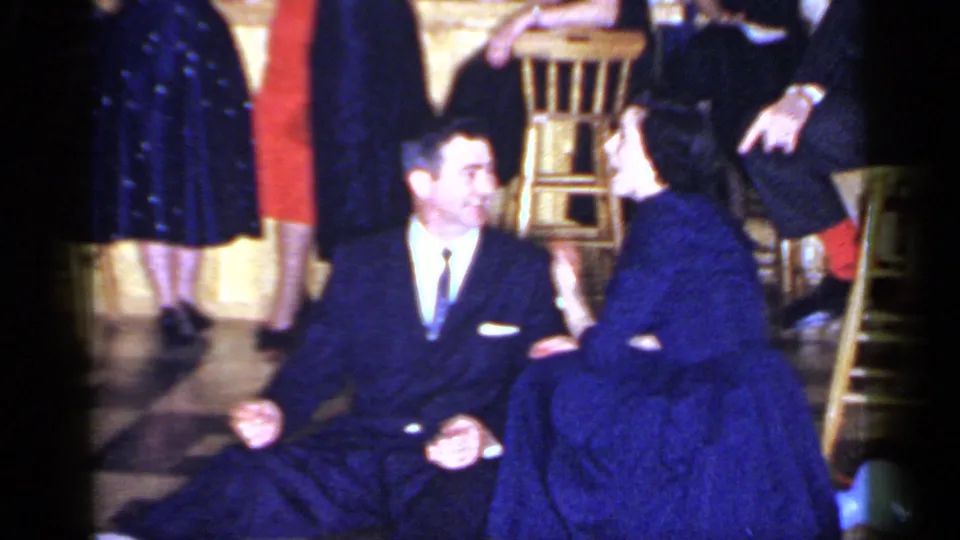The couple sits close together on the Mineola dance floor, surrounded by guests in formal clothing and wooden stools.