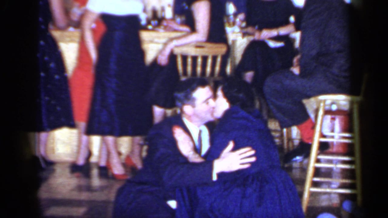 The suited man and woman in a blue dress kiss on the floor during a 1966 Mineola social gathering, with red heels and guests visible nearby.