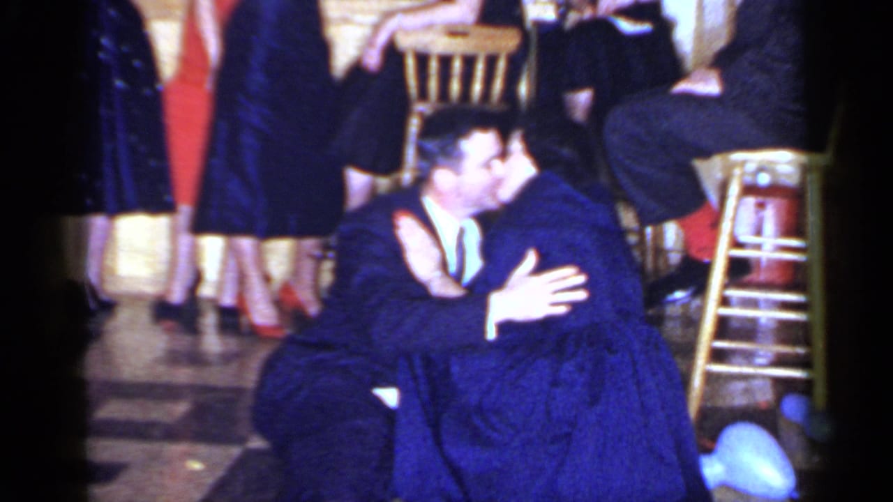 The couple continues kissing and embracing on the Mineola dance floor, framed by formal guests, stools and the warm 8mm home-movie look.