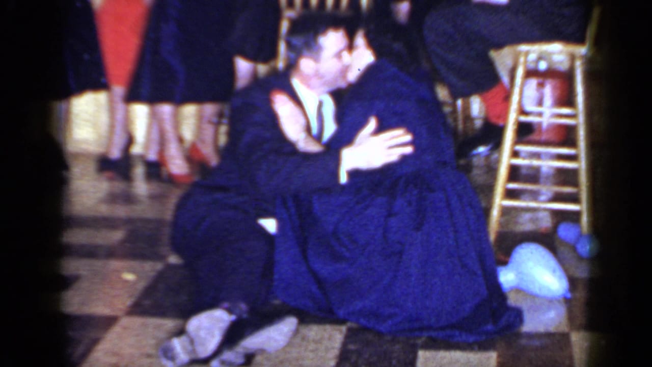 Man in a dark suit embraces a woman in a blue dress on a checkered floor.