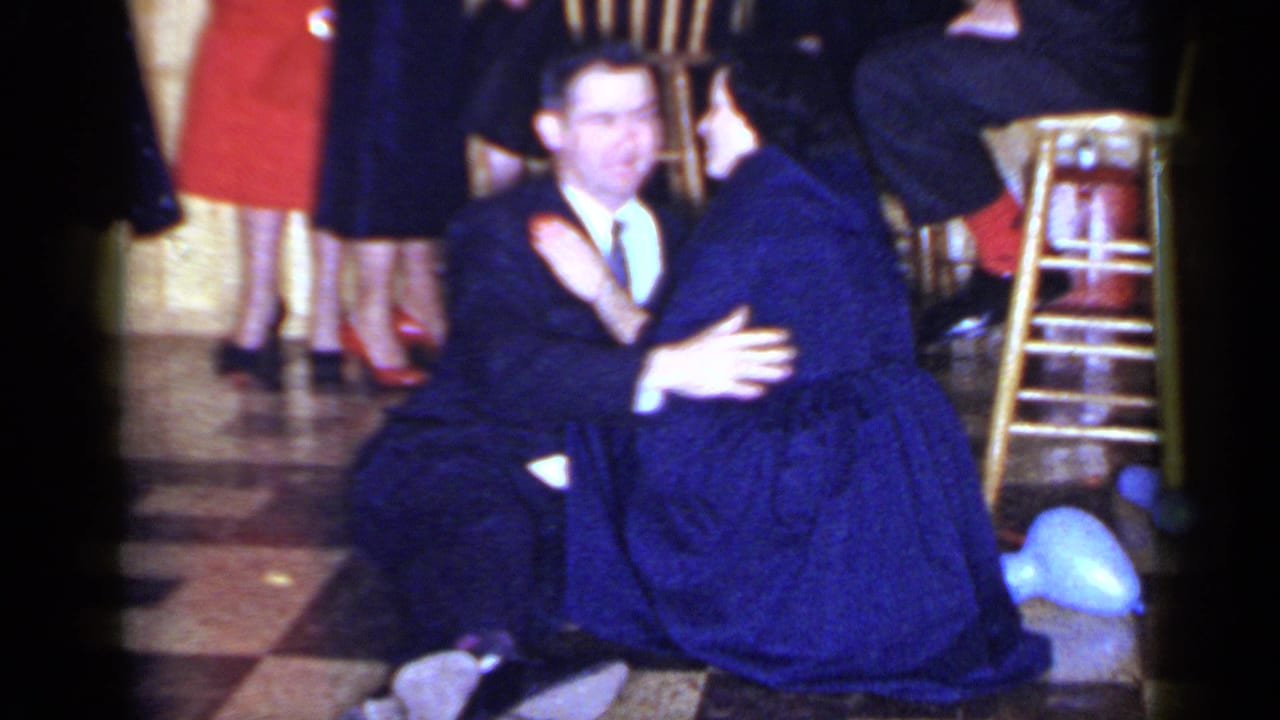 Suited man kneels beside a seated woman in a blue dress near stools and balloons.
