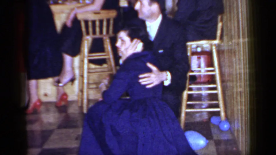 The home-movie frame shows the woman seated on the checkered floor with the suited man close behind her at the 1966 Mineola event.