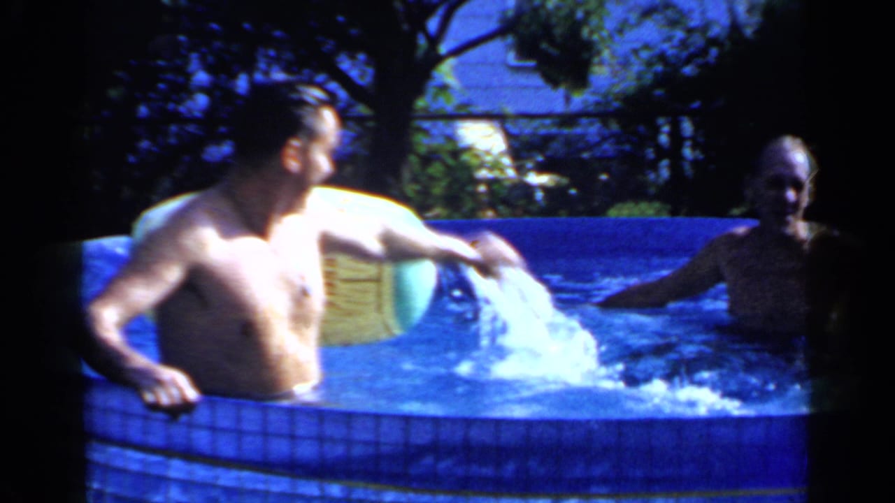 1966 Mineola frame of adult men in a backyard pool, one creating a splash beside a pale inflatable ball near the pool wall.