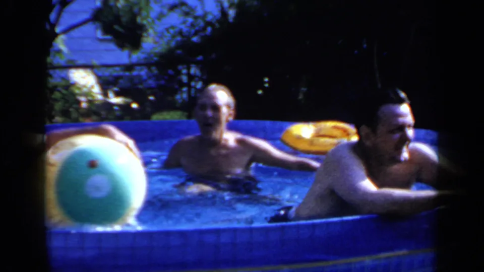 1966 Mineola home-movie frame showing two adult men in a blue above-ground pool, with a large beach ball and shaded yard behind them.