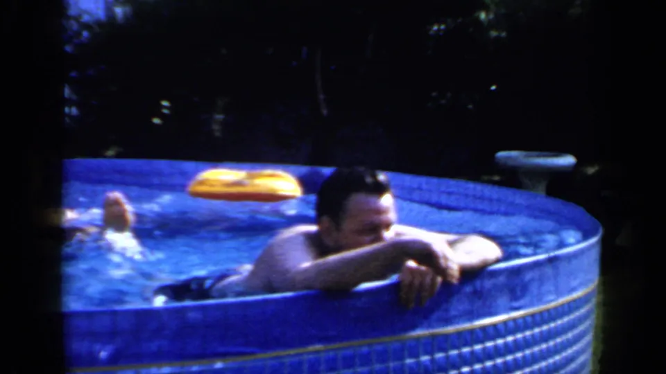 1966 Mineola frame of an adult man leaning on the edge of a backyard above-ground pool, with a yellow float behind him.