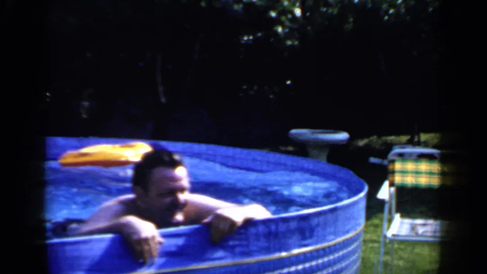 In 1966 Mineola home-movie footage, an adult man leans on the edge of a blue backyard pool with a yellow inflatable behind him.