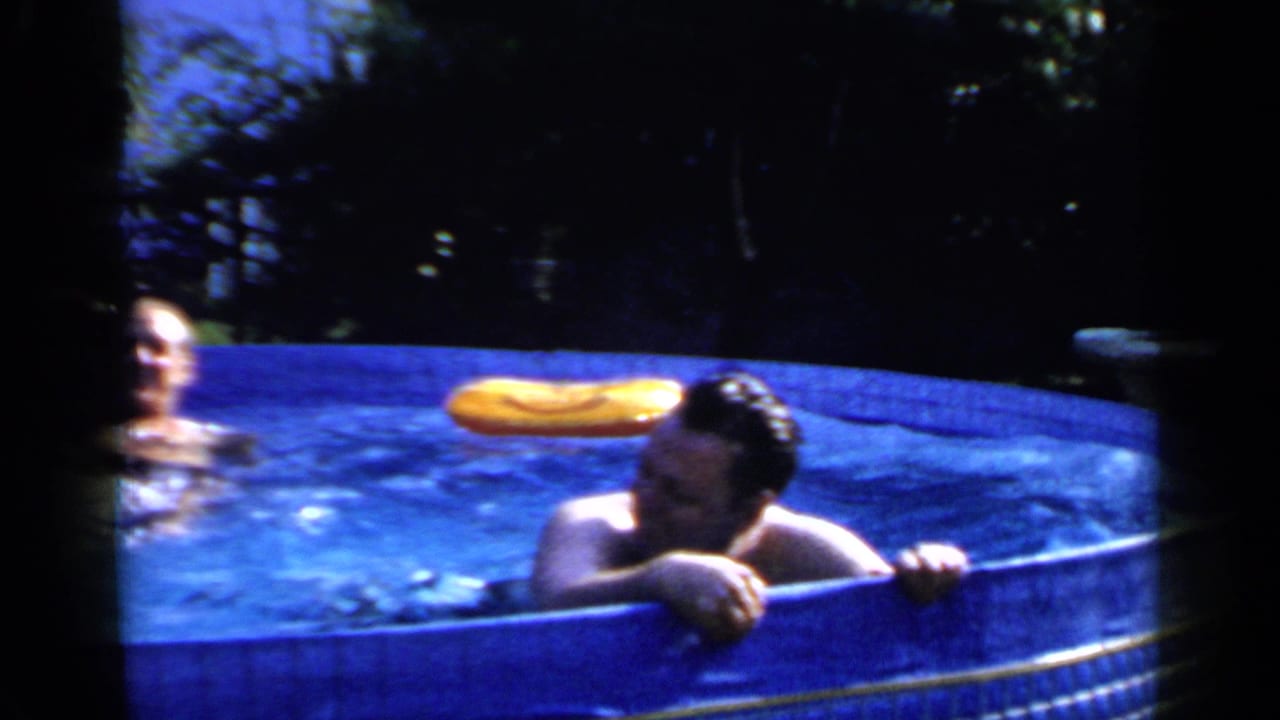 An adult man leans against a blue backyard pool while another man is partly visible in the water behind him in Mineola, New York.
