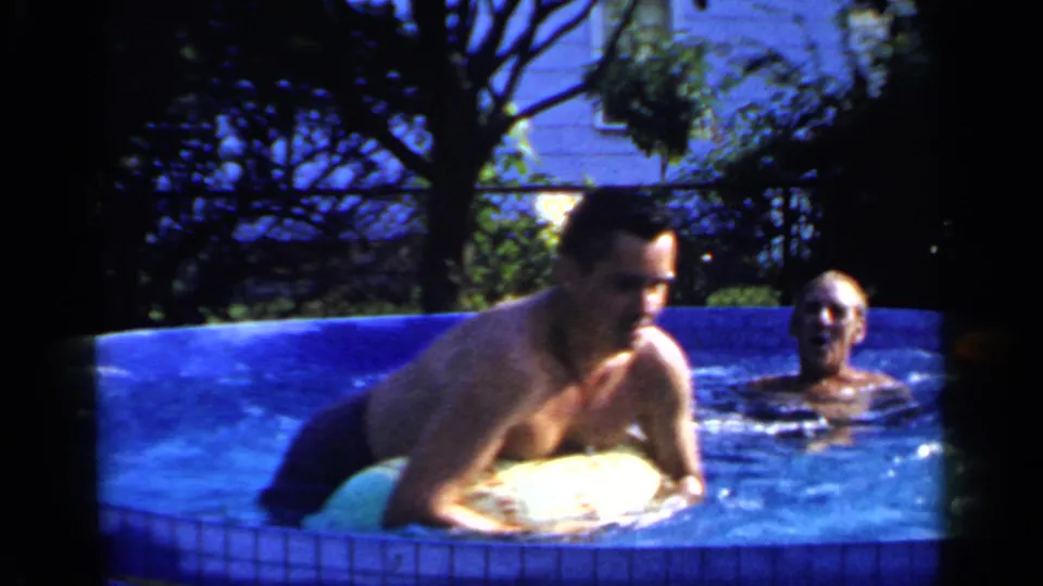 A man leans over a yellow inflatable ring at the edge of the blue backyard pool while another man is visible behind him.
