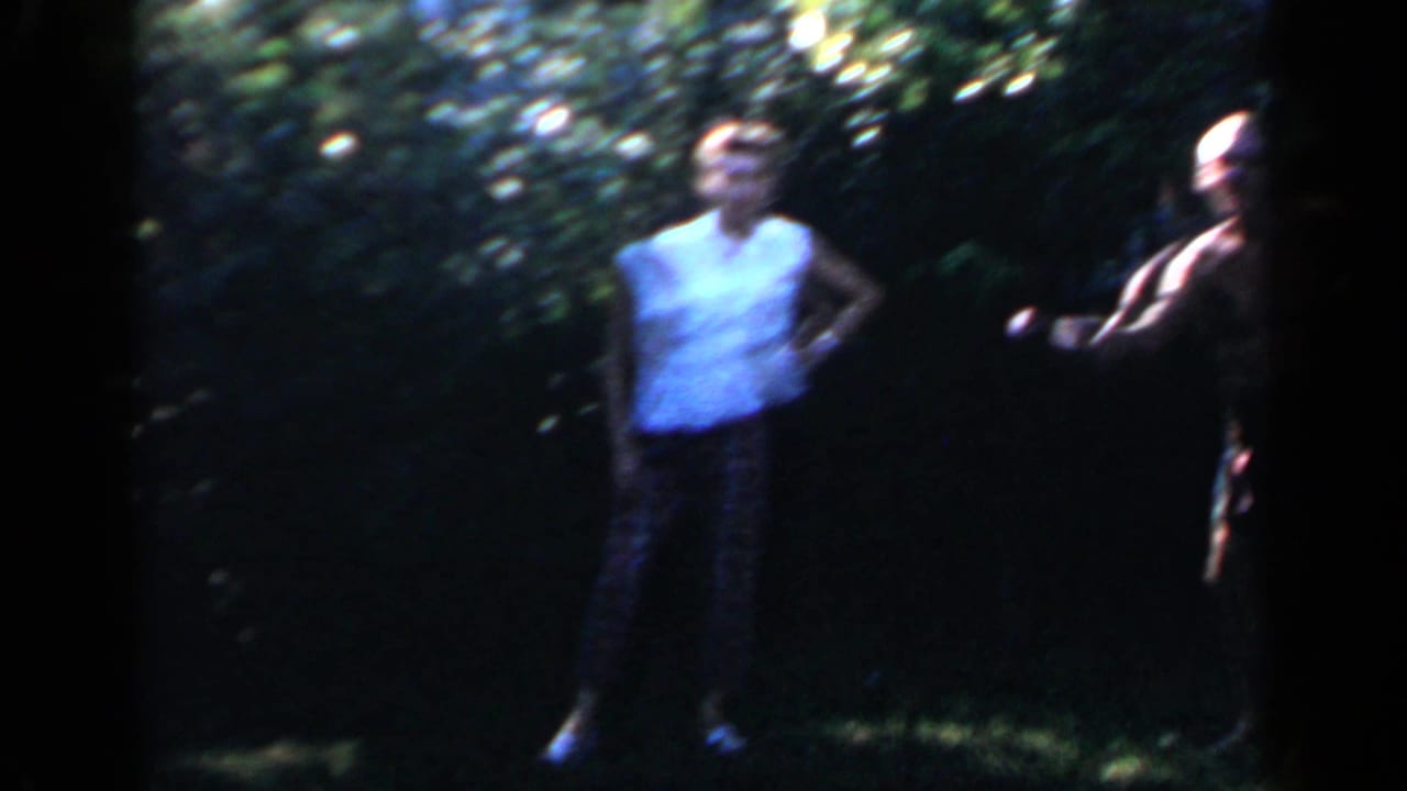 In 1966 Mineola home-movie footage, two adults stand on a shaded backyard lawn during a casual outdoor gathering.