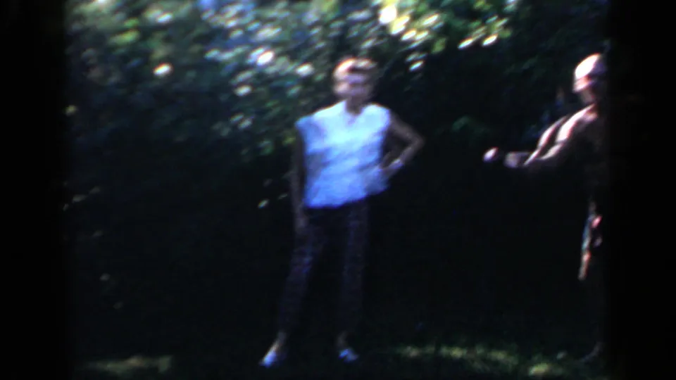 In 1966 Mineola home-movie footage, two adults stand on a shaded backyard lawn during a casual outdoor gathering.