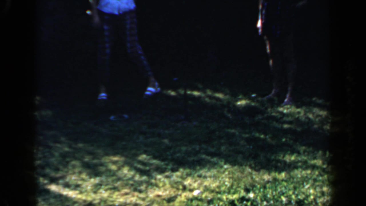 A partial 1966 Mineola backyard view shows people standing on grass, with legs and shoes visible in mixed sunlight and shade.