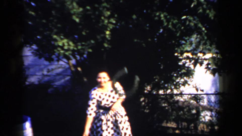 A woman in a polka-dot dress stands near foliage and a fence in the 1966 Mineola backyard home-movie scene.
