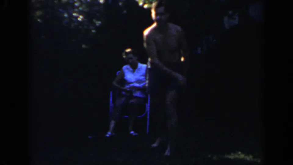 In the dim 1966 Mineola backyard footage, one adult sits in a folding chair while another stands nearby on the grass.