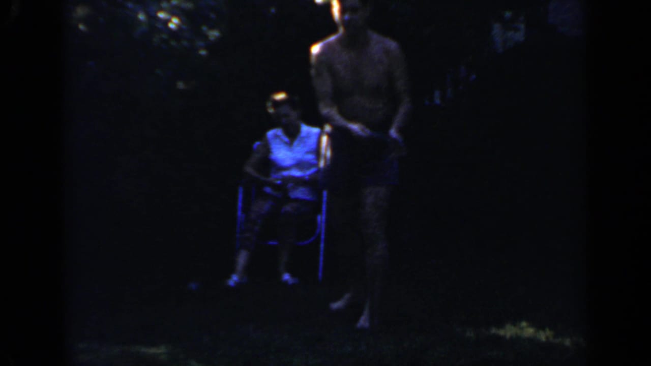 Man in blue shorts stands near a seated woman in a dark backyard.