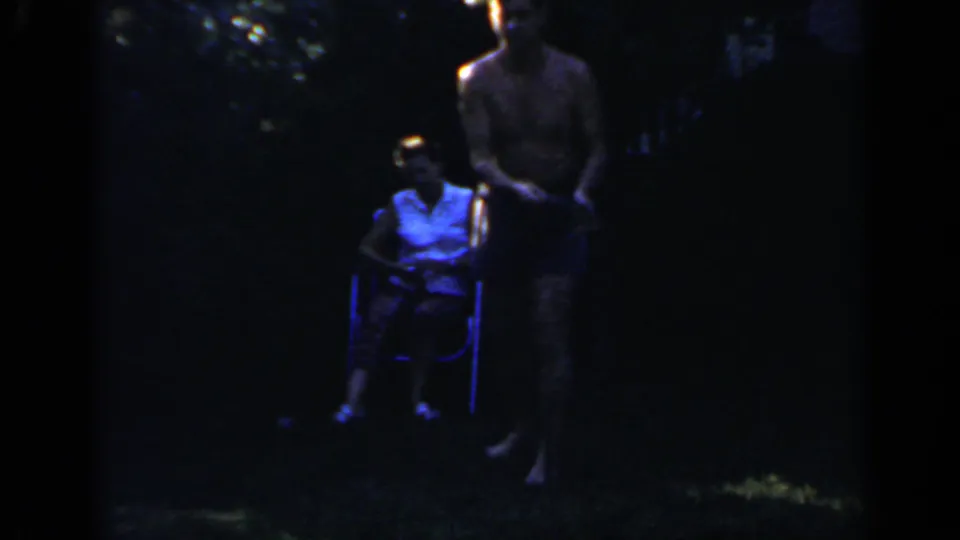 In 1966 Mineola home-movie footage, a man in blue shorts stands by a woman seated in a folding chair in a dim backyard.