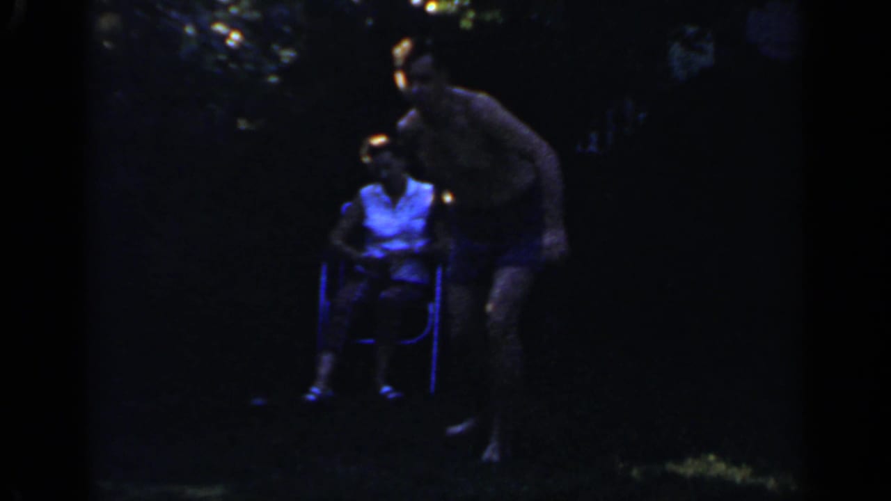 Man bends forward beside a seated woman in a dark outdoor yard.