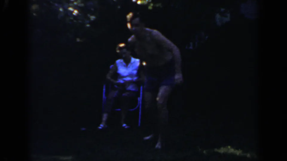 The man in blue shorts leans forward near a woman in a folding chair during the Mineola backyard horseshoe scene.