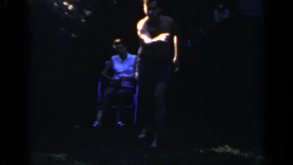A shirtless man passes through the foreground while a seated woman remains visible in the dark Mineola backyard.
