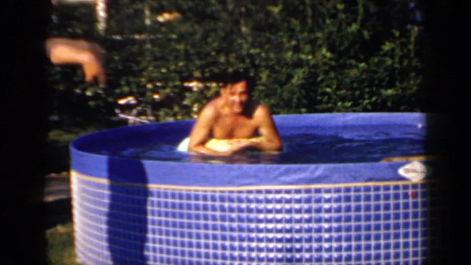 Adult man in a blue backyard pool rests on a yellow float in sunny 1966 Mineola home-movie footage.