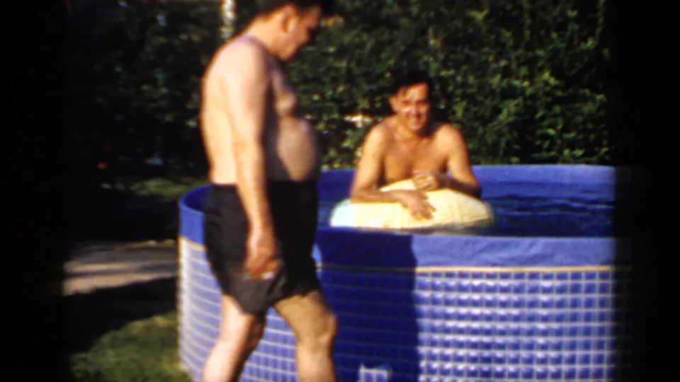 Two adult men appear beside a blue backyard pool, one standing in black trunks and one seated on a yellow float.