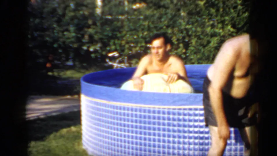 Adult man in a blue backyard pool holds a yellow float while another man in dark trunks leans at the edge.