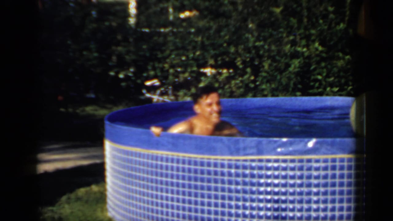 Smiling adult man in a blue inflatable pool with greenery behind him.