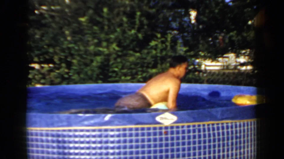 A man floats belly-down across a large ball in the blue backyard pool, with an orange inner tube near the far edge.