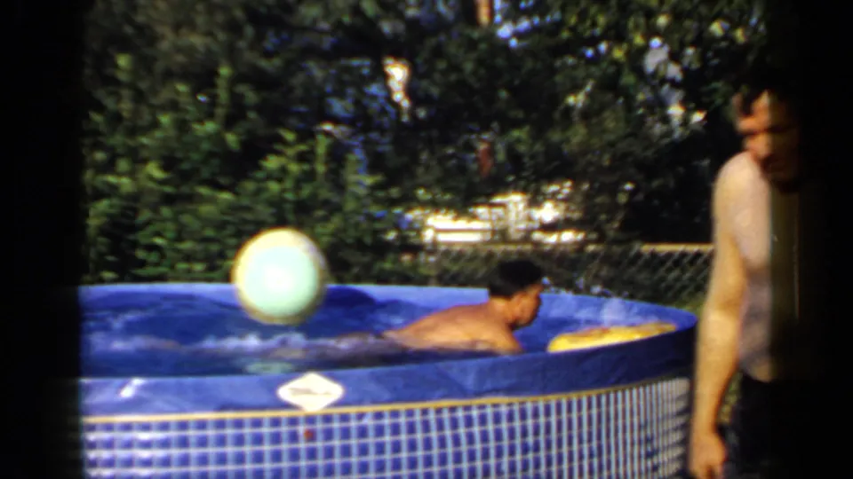 An adult floats in the blue backyard pool near a beach ball while another adult in dark swim trunks stands at the edge.