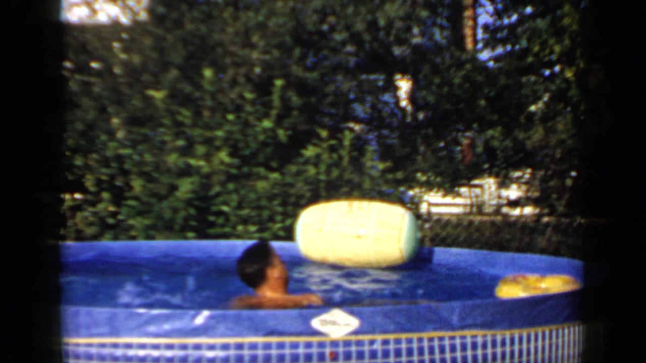 Man seated in a blue inflatable pool with a yellow float and trees behind him.