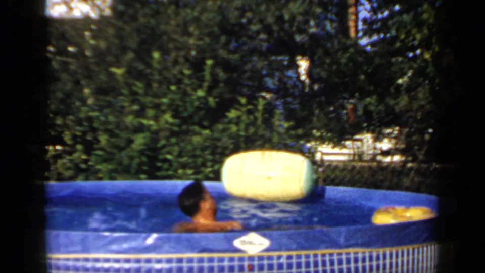 1966 Mineola home-movie frame showing a man relaxing in a blue backyard inflatable pool with trees and a yellow float nearby.