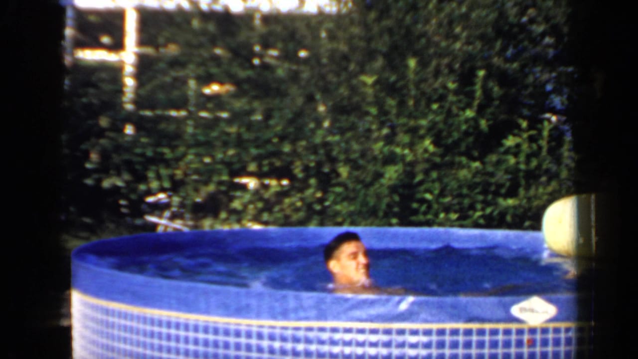 Man standing or floating in a blue inflatable pool near a pale cushion and trees.