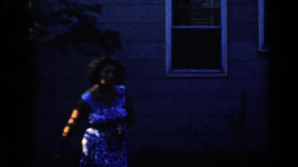 1966 Mineola home-movie frame showing a woman in a blue patterned dress near blue house siding and a window in shadow.