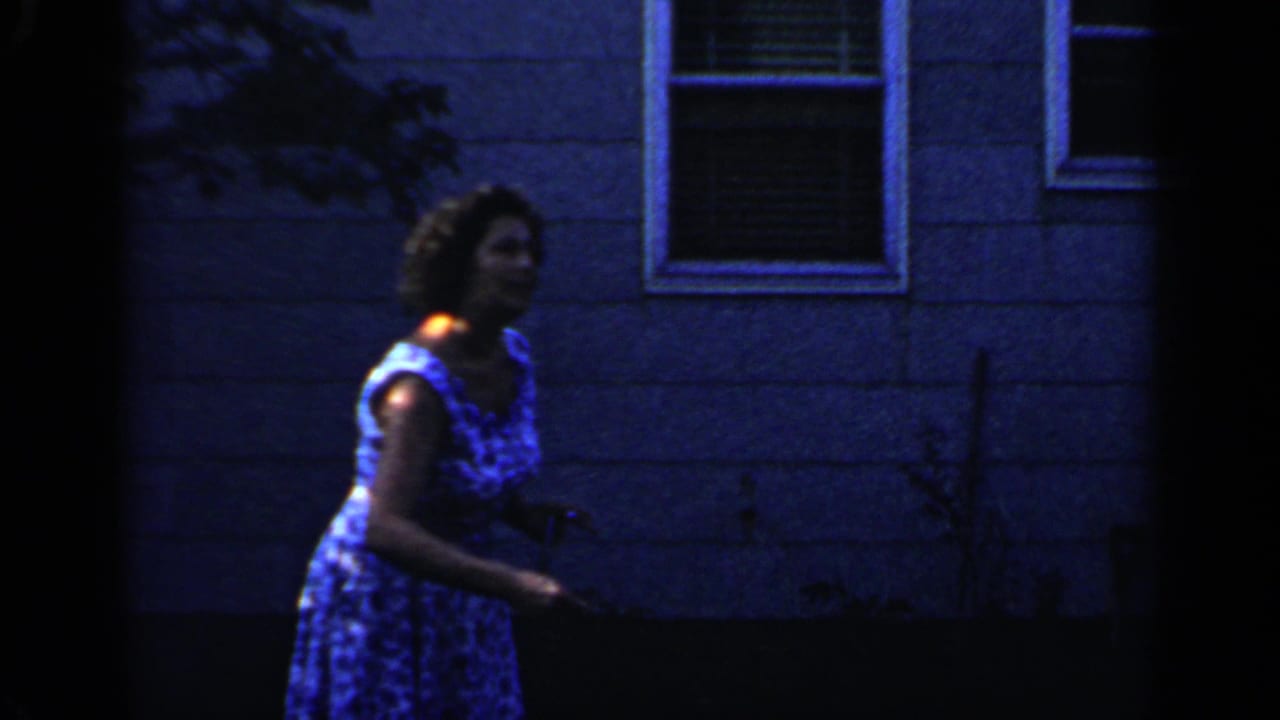 Woman in a blue patterned dress standing near a house wall in a shaded backyard.