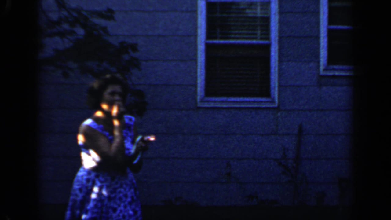 The woman raises a small lighted object near her face in the Mineola backyard, with the house wall and window behind her.