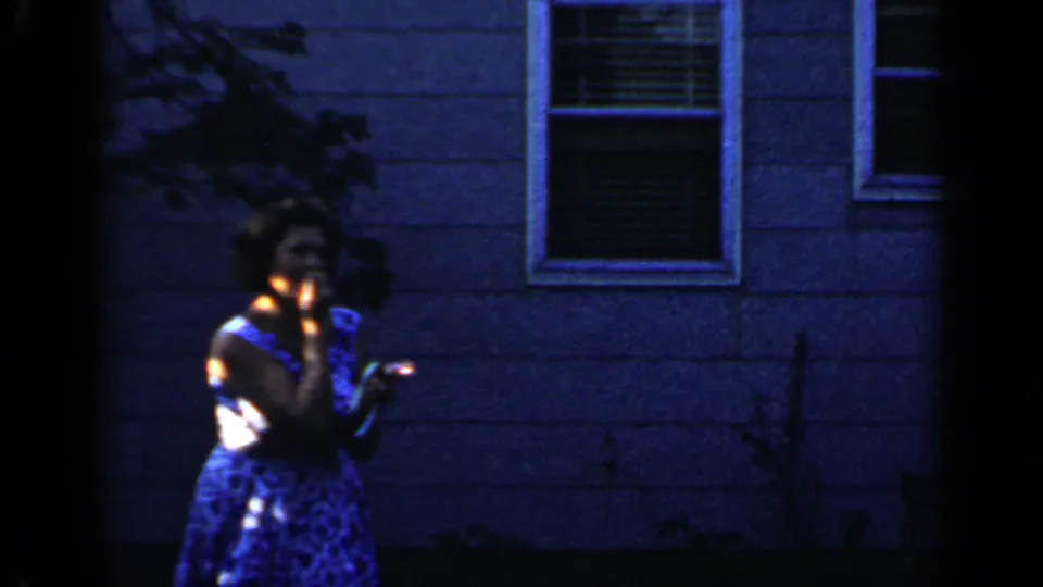 The woman raises a small lighted object near her face in the Mineola backyard, with the house wall and window behind her.