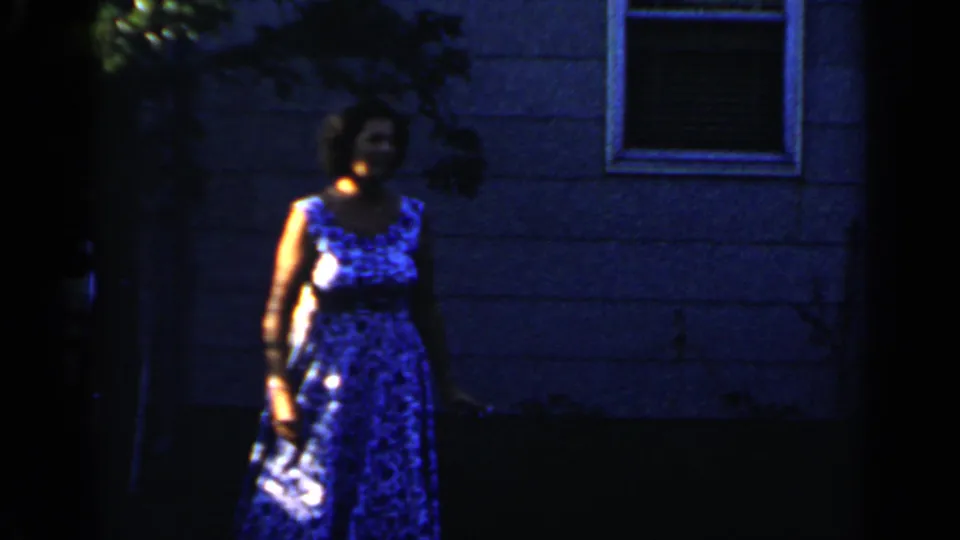 A woman in a blue patterned dress faces the handheld camera in a dark 1966 Mineola backyard scene.
