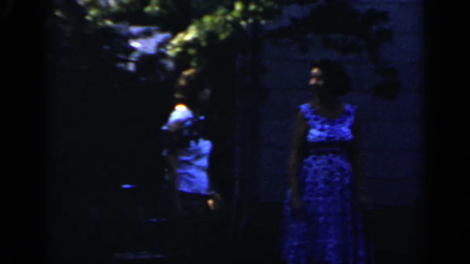 The woman stands at the edge of the shaded yard while another person briefly appears in the background of the Mineola home movie.