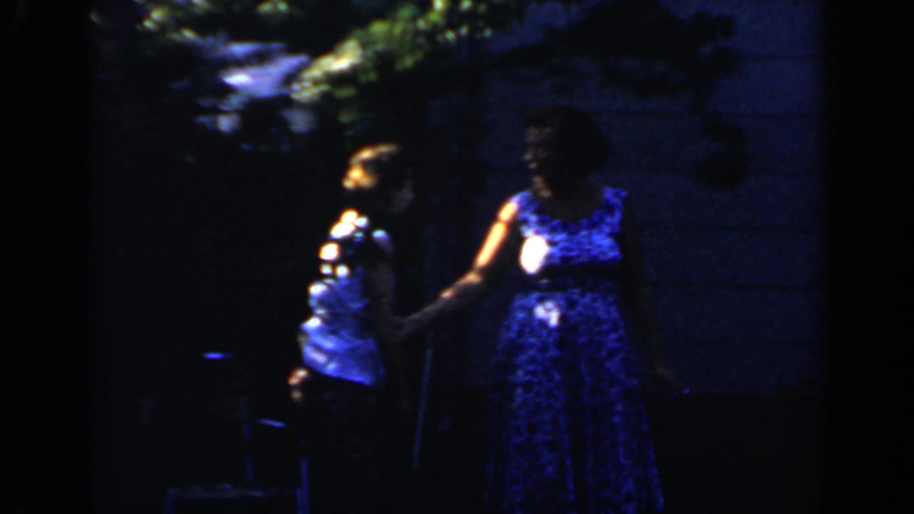 Two women in summer clothing stand in a dark backyard near a house exterior.