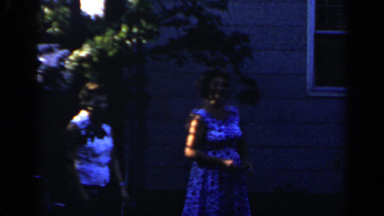 Two women stand apart in deep shade near a house wall and shrubs.