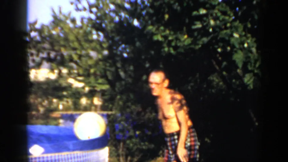1966 home-movie frame of a shirtless man in plaid swim trunks beside a small blue pool in a leafy yard.