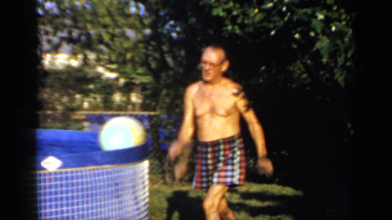 Shirtless man in plaid swim trunks stands near a blue above-ground pool in a sunny backyard.