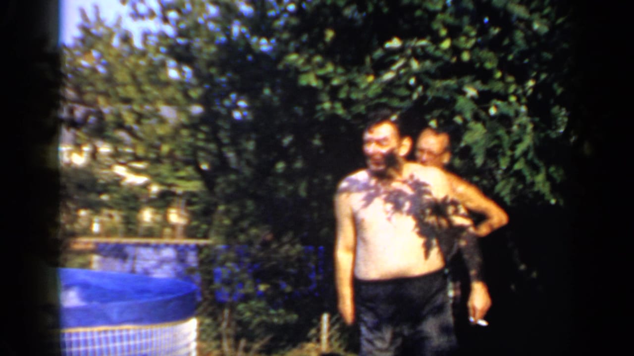 Man in swim trunks laughs near a blue above-ground pool while another adult stands behind him.