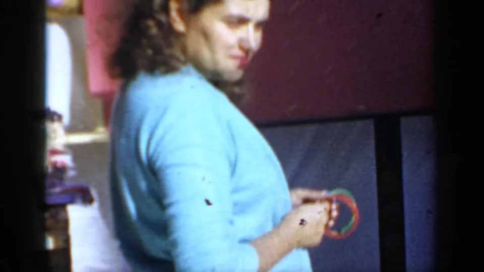 A woman in a light blue blouse stands near a red wall in the 1966 Mineola backyard scene, looking toward the side of frame.