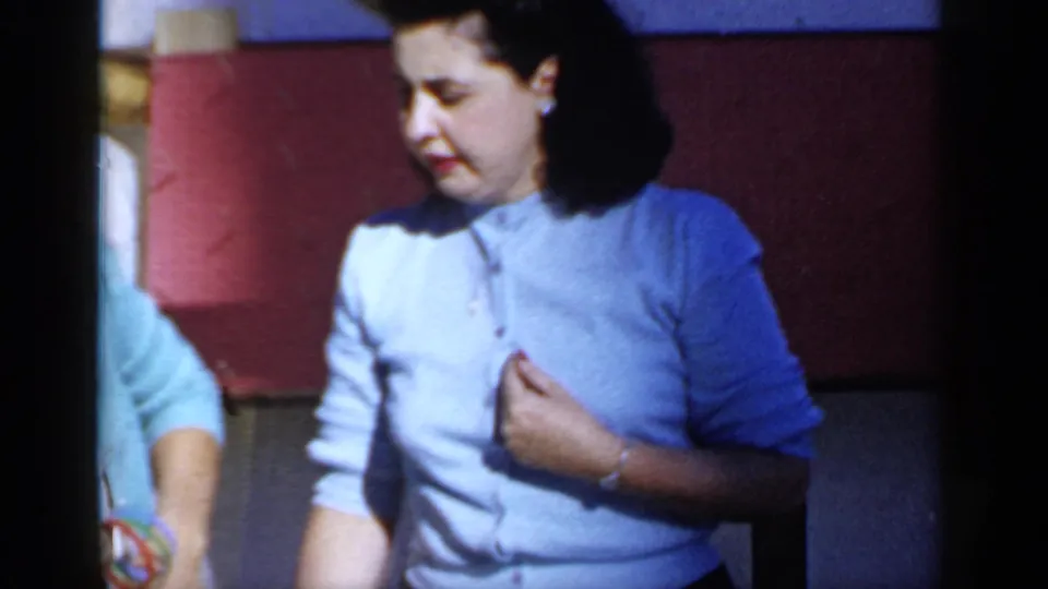 A woman in a pale blue cardigan or blouse stands in the Mineola yard footage, framed closely against a red house wall.